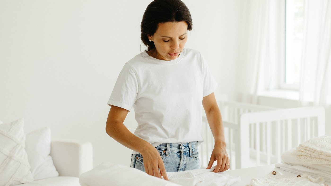 Vânia folding a small baby garment at a white table, a crib behind her