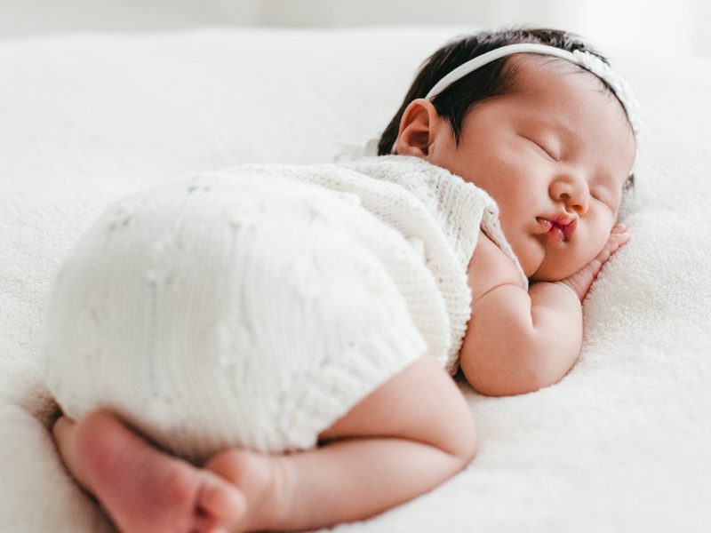 A newborn in a white knit romper asleep on a white blanket